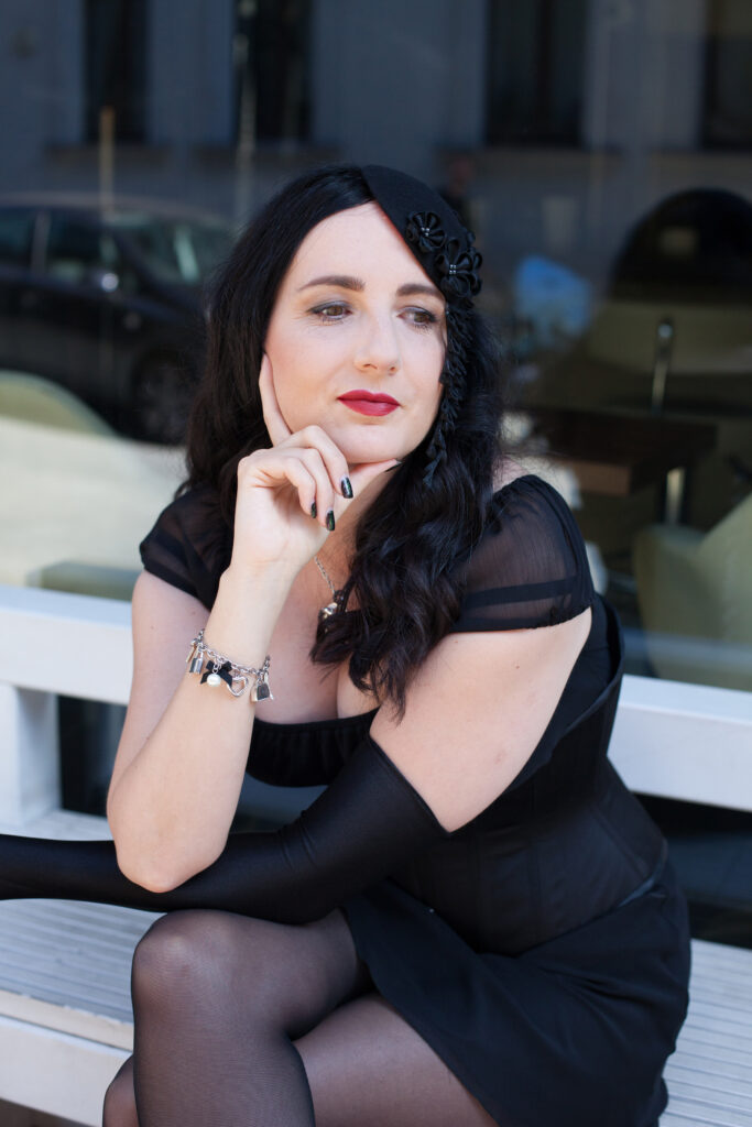 Jennifer Sonntag sitzt auf einer weißen Bank vor einem Schaufenster. Sie trägt ein kurzes schwarzes Kleid mit einer schwarzen Corsage. Ihr linker Arm ist mit einem langen schwarzen Handschuh bekleidet. Ihr Kinn hat sie sinnierend auf ihre rechte Hand gestützt. Sie hat die Beine übereinander geschlagen. Ihre langen schwarzen Haare trägt sie offen und mit einem schwarzen Faszinator (kleiner Hut zum in die Haare stecken) verziert.